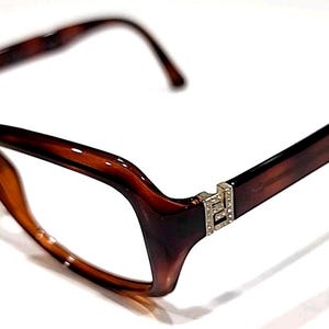 Può includere: Occhiali marroni con montatura tartarugata di forma rettangolare. Gli occhiali hanno un accento decorativo argentato sulle aste. Le lenti sono trasparenti. Gli occhiali sono su uno sfondo bianco.