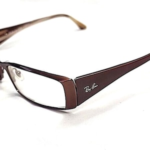 Puede incluir: Gafas Ray-Ban marrones con lentes rectangulares. Las monturas son de color marrón oscuro con un sutil degradado en las patillas. El logotipo de Ray-Ban es visible en la patilla derecha. Las gafas están sobre un fondo blanco.