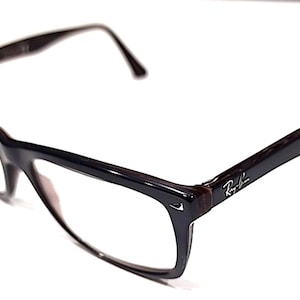 May include: Dark brown Ray-Ban eyeglasses with a rectangular frame. The glasses have a glossy finish and the Ray-Ban logo is visible on the temple. The lenses are clear.