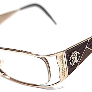 May include: Gold-toned eyeglasses with a rectangular frame and a geometric design on the temples. The temples feature a dark brown and light brown pattern with the "Roberto Cavalli" logo. The arms are black.