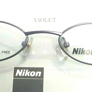 Puede incluir: Gafas Nikon de titanio violeta oscuro de forma ovalada. Las monturas finas muestran el texto "TITANIUM NICKEL FREE" y "Nikon titanium". Las gafas tienen un diseño minimalista y son adecuadas para el uso diario.