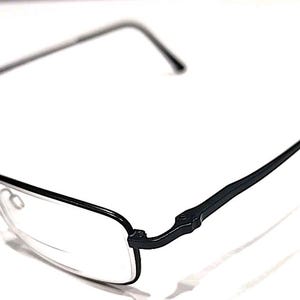 Puede incluir: Gafas rectangulares negras con montura metálica fina y lentes transparentes. Las patillas también son negras y tienen una ligera curva. Las gafas están sobre un fondo blanco.