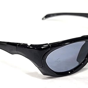 May include: Black sunglasses with grey lenses. The sunglasses have a sporty design with thick, curved frames and arms. The lenses are oval-shaped and provide a contrast to the black frames. The sunglasses are set against a white background.