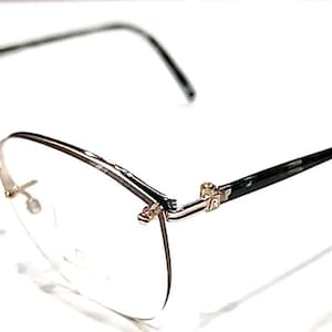 Op de afbeelding: Bril zonder montuur met een goudkleurig metalen montuur en heldere glazen. De pootjes zijn zwart met een subtiel patroon. De bril staat tegen een witte achtergrond, wat het delicate ontwerp benadrukt.