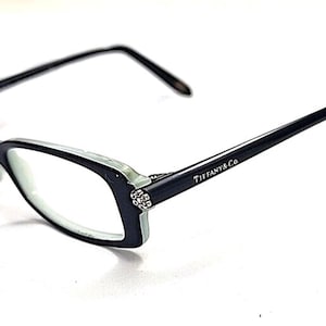 May include: Black and light blue eyeglasses with the Tiffany & Co. logo on the temple. The frames have a rectangular shape with a decorative embellishment on the corner. The glasses are set against a white background.