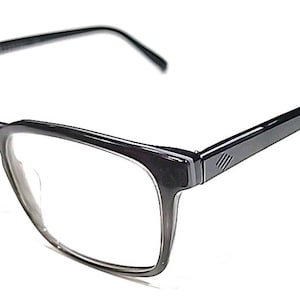 May include: Dark grey rectangular eyeglasses with clear lenses. The frames have a thick, matte finish. The temples feature a minimalist design with three parallel lines. The glasses are set against a white background. The overall design is modern.