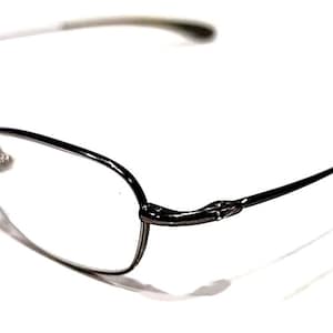 Op de afbeelding: Een bril met een dun, zwart metalen montuur en heldere glazen. De pootjes zijn slank en buigen zachtjes naar het oor. De bril staat tegen een heldere witte achtergrond.