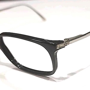 Puede incluir: Gafas negras con montura rectangular y varillas plateadas. Las monturas tienen un acabado brillante y pequeños detalles decorativos cerca de los cristales. Estas gafas son para la corrección de la visión.