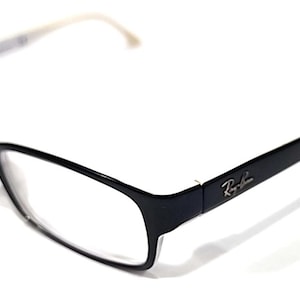 Puede incluir: Gafas rectangulares negras con lentes transparentes y una patilla blanca y plateada. El logotipo de Ray-Ban es visible en la patilla. Las gafas están sobre un fondo blanco.