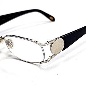 May include: Silver-framed spectacles with oval lenses and black arms. The spectacles have a distinctive design with circular silver accents on the arms. The lenses are clear, and the overall style is modern and sleek.