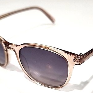 May include: A pair of sunglasses with a light brown frame and dark gray lenses. The sunglasses have a classic rectangular shape and are set against a white background. The arms of the sunglasses are thin and extend back.