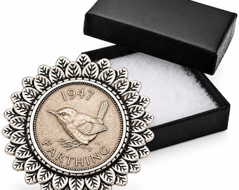 Peut inclure: Une broche argentée présentant une pièce de farthing de 1947 au centre, entourée d'un cadre décoratif de feuilles en relief. La broche est présentée à côté d'une boîte cadeau rectangulaire noire ouverte avec une doublure blanche.