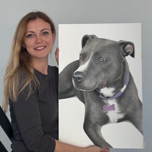 May include: A portrait of a gray dog with a white patch on its face, wearing a purple collar and a tag that says "Eve". The artwork is held by a person with long blonde hair.