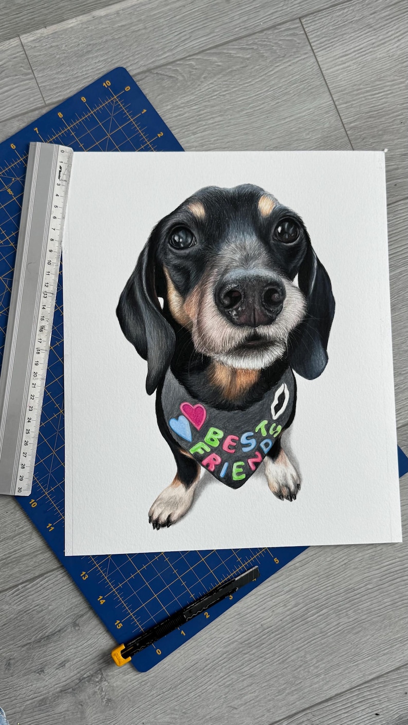May include: A detailed watercolor painting of a Dachshund dog with a black and tan coat, wearing a bandana that says "BEST FRIEND" in colorful letters. The dog is centered on a white background, with a blue cutting mat and ruler in the background.