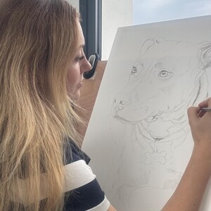 May include: A pencil drawing of a dog is being created on a white canvas. The artist is wearing a navy blue and white striped shirt. The dog portrait is detailed, with a bone-shaped tag visible.