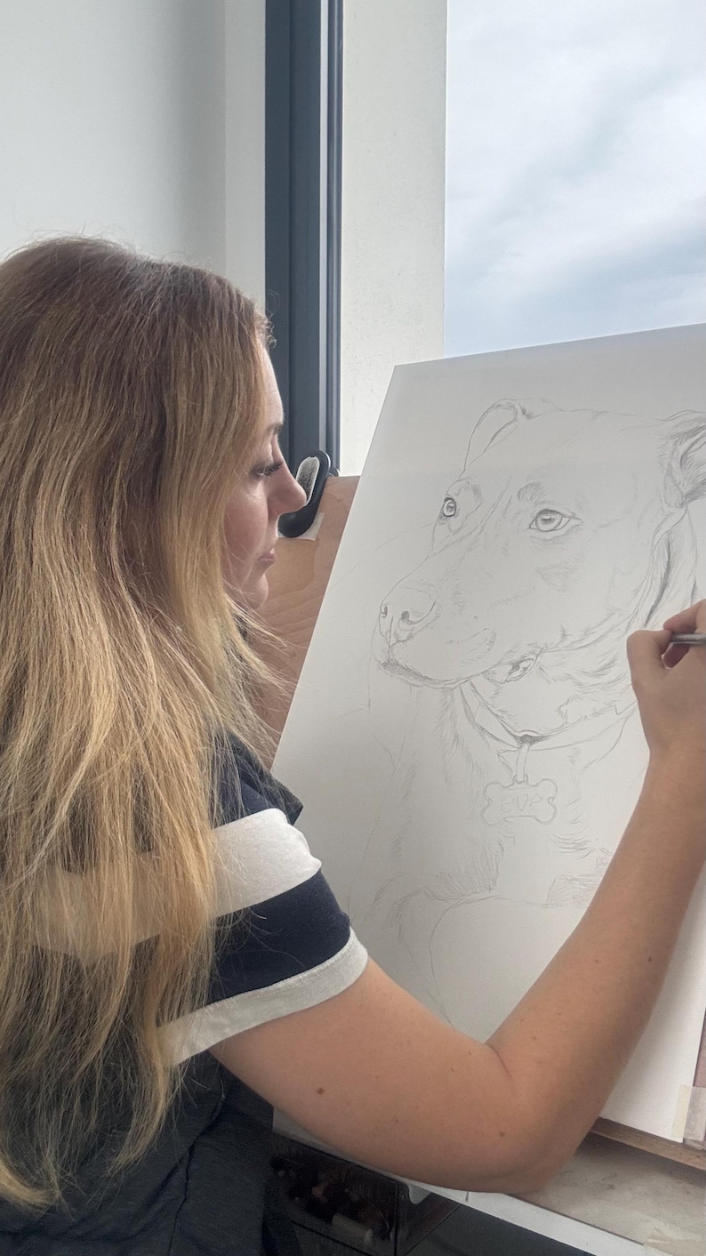 May include: A pencil drawing of a dog is being created on a white canvas. The artist is wearing a navy blue and white striped shirt. The dog portrait is detailed, with a bone-shaped tag visible.
