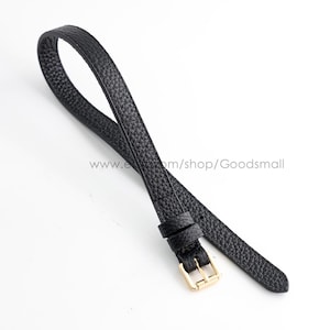May include: A black watch strap with a textured, pebbled surface finish. The strap features a gold-toned metal buckle and two matching black leather loops to secure the end. The strap is looped over itself, showing the exterior and interior texture.