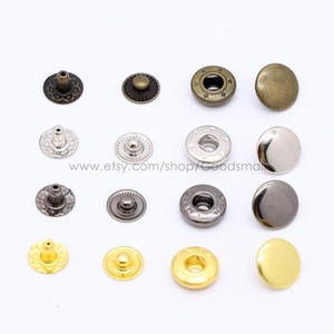 May include: A collection of sixteen metal snap fasteners arranged in four rows and four columns. The set includes bronze, silver, gunmetal, and gold finishes, featuring various circular designs, smooth caps, and functional stud and socket components.