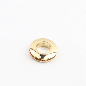 May include: A gold-colored metal grommet with a smooth, shiny finish. The grommet has a circular shape with a hole in the center.