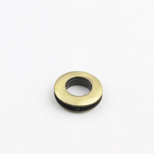 May include: A gold metal grommet with a black inner ring. The grommet is a circular shape with a hole in the center.