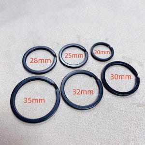 Puede incluir: Se muestran seis llaveros negros de diferentes tamaños. Los anillos varían de 20 mm a 35 mm de diámetro. Cada anillo tiene su tamaño impreso en rojo. Los llaveros están dispuestos sobre una superficie gris claro.