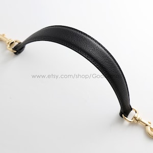 Puede incluir: Un asa de bolso negra arqueada con superficie texturizada y bordes cosidos. El asa cuenta con un mosquetón giratorio de metal dorado pulido en cada extremo, lo que permite una fácil conexión a un bolso o accesorio.