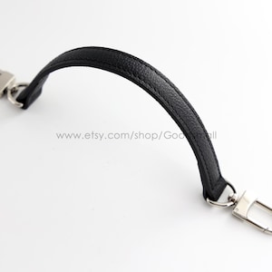 Puede incluir: Una correa de muñeca de cuero negro texturizado con mosquetones giratorios de metal plateado en ambos extremos. La correa tiene una forma curva y arqueada e incluye el texto www.etsy.com/shop/Goodsmall impreso en el centro.