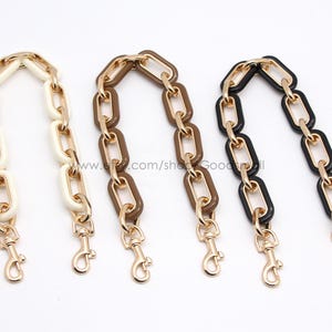 May include: Three handbag chain straps in ivory, brown, and black, each with gold-tone metal links and lobster claw clasps. The chains are displayed against a white background, showcasing their colour variations.