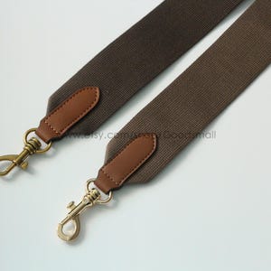 May include: Two brown canvas bag straps with leather accents and gold-colored metal clasps. The straps are angled across a white background, showcasing their design and hardware.