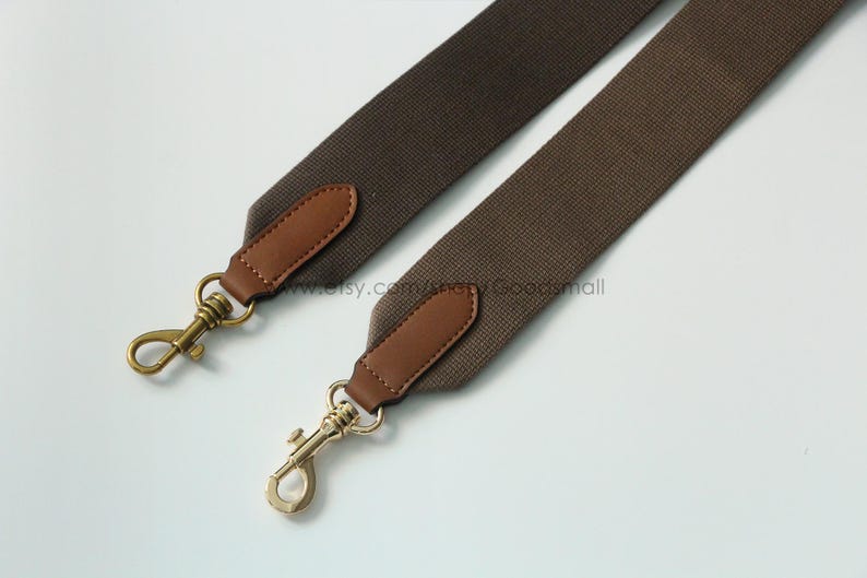 May include: Two brown canvas bag straps with leather accents and gold-colored metal clasps. The straps are angled across a white background, showcasing their design and hardware.