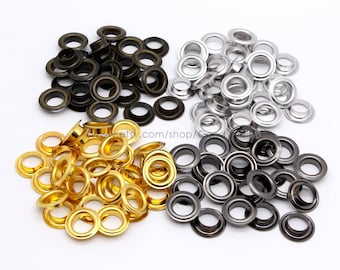 May include: Assortment of metal eyelets in gold, silver, and black. The eyelets are arranged in three piles on a white surface.