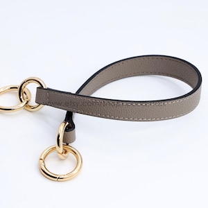 Puede incluir: Una correa de muñeca de cuero color topo con superficie texturizada y bordes negros. La correa está unida a dos anillos de metal dorado pulido, con un anillo conectado a un cierre circular de resorte más pequeño para una fijación segura.