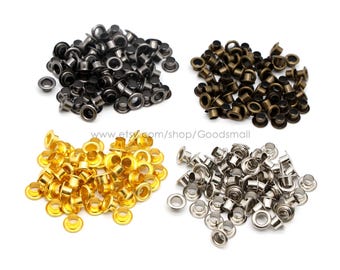 May include: Four piles of metal eyelets in different finishes: gold, silver, black, and bronze. The eyelets are small, round, and have a hole in the center. They are used for attaching fabric or other materials together.