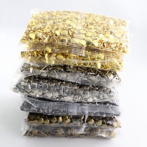May include: A vertical stack of five clear plastic bags filled with small metal rivets. The top bag contains gold-colored rivets, followed by bags of silver, gunmetal, and bronze-toned rivets, each featuring a smooth, rounded cap design.