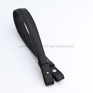 Puede incluir: Una correa de muñeca de cuero negro texturizado con diseño de lazo, que cuenta con un pequeño cierre de perno de metal plateado y una trabilla deslizante a juego para asegurar la correa en su lugar, sobre un fondo blanco liso.
