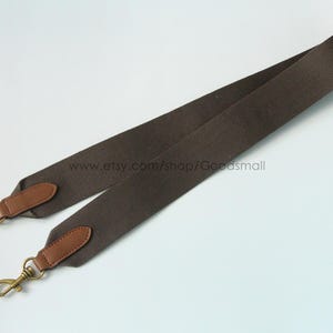 May include: A brown shoulder bag strap with leather accents and gold-colored metal clasps. The strap is made of a durable, woven material. The strap is designed to be attached to a bag.