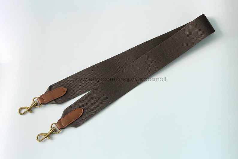 May include: A brown shoulder bag strap with leather accents and gold-colored metal clasps. The strap is made of a durable, woven material. The strap is designed to be attached to a bag.