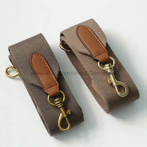 May include: Two canvas bag straps in a neutral brown color, each featuring a brown leather accent and gold-colored metal hardware. The straps have a classic design with a hook closure.