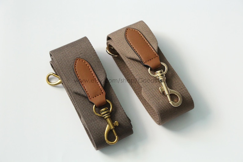 May include: Two canvas bag straps in a neutral brown color, each featuring a brown leather accent and gold-colored metal hardware. The straps have a classic design with a hook closure.