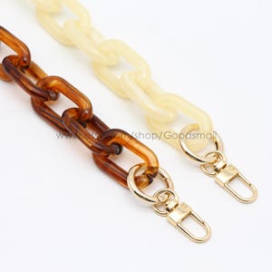 Puede incluir: Dos correas de cadena de plástico marrón y crema con cierres de metal dorado. Las correas están unidas y probablemente sean para un bolso de mano o una cartera.