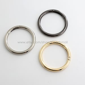 May include: Three circular metal spring rings in silver, gunmetal, and gold tones. Each ring features a small hinge mechanism for opening and closing. The text www.etsy.com/shop/Goodsmall is printed in the center of the three metal rings.