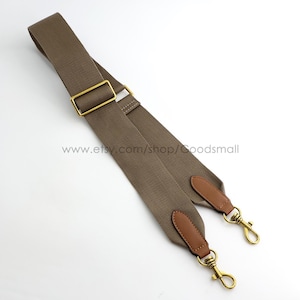 May include: A taupe-colored adjustable bag strap made of woven fabric, featuring brass-toned metal hardware, including a rectangular slider and two swivel lobster claw clasps. The strap ends are reinforced with brown leather tabs.