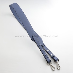 May include: A periwinkle blue bag strap featuring a textured surface, adjustable length settings with silver-toned metal studs, and two silver-toned swivel lobster claw clasps at the ends for secure attachment to a handbag or accessory.