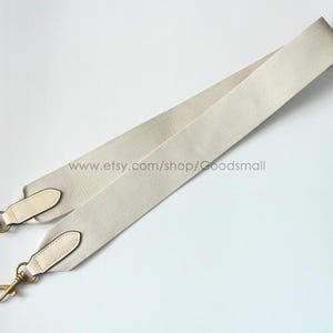 May include: Cream-colored bag strap with gold-tone hardware. The strap is made of a ribbed fabric and features leather accents at the ends. The strap has two gold-tone clasps for attachment. The Etsy shop URL is visible.