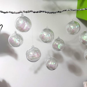 Op de afbeelding: Een horizontale stang van gedraaid zilverkleurig draad waaraan acht doorzichtige bolvormige ornamenten met een iriserende binnenkant hangen. Links hangt een aparte verticale ketting van kleine, veelkleurige kralen aan de draad. De ornamenten hangen op verschillende hoogtes.