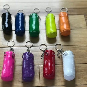 Pode incluir: Uma coleção de chaveiros cilíndricos coloridos, em preto, azul, verde, amarelo, laranja, rosa, roxo, vermelho e branco. Cada chaveiro tem uma corrente e um anel prateados.