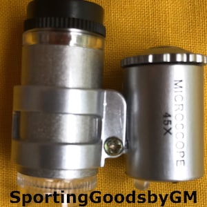 Puede incluir: Un microscopio de mano plateado y negro con un aumento de 45x. El microscopio tiene una luz LED incorporada y una perilla de ajuste de enfoque. El texto "MICROSCOPE 45X" está impreso en el lateral del microscopio.