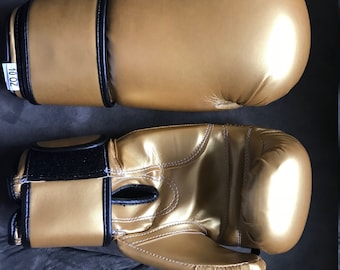 May include: A pair of gold boxing gloves with black trim. The gloves have a white stitching detail and the text "700L" is visible on one of the gloves.