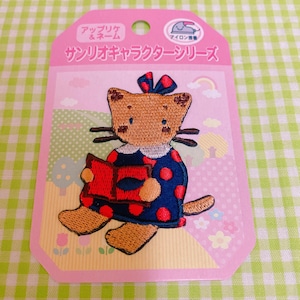 May include: An iron-on patch featuring a brown cat wearing a blue and red polka dot dress and holding a red book. The patch is surrounded by a pink background with the text "サッリキキャラクターシリーズ" in Japanese.