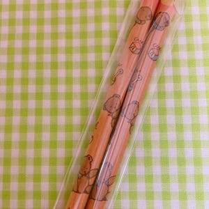May include: Two brown wooden pencils with a clear plastic wrapper. The pencils are decorated with a repeating pattern of brown bears and colorful blocks. The pencils are on a green and white checkered background.